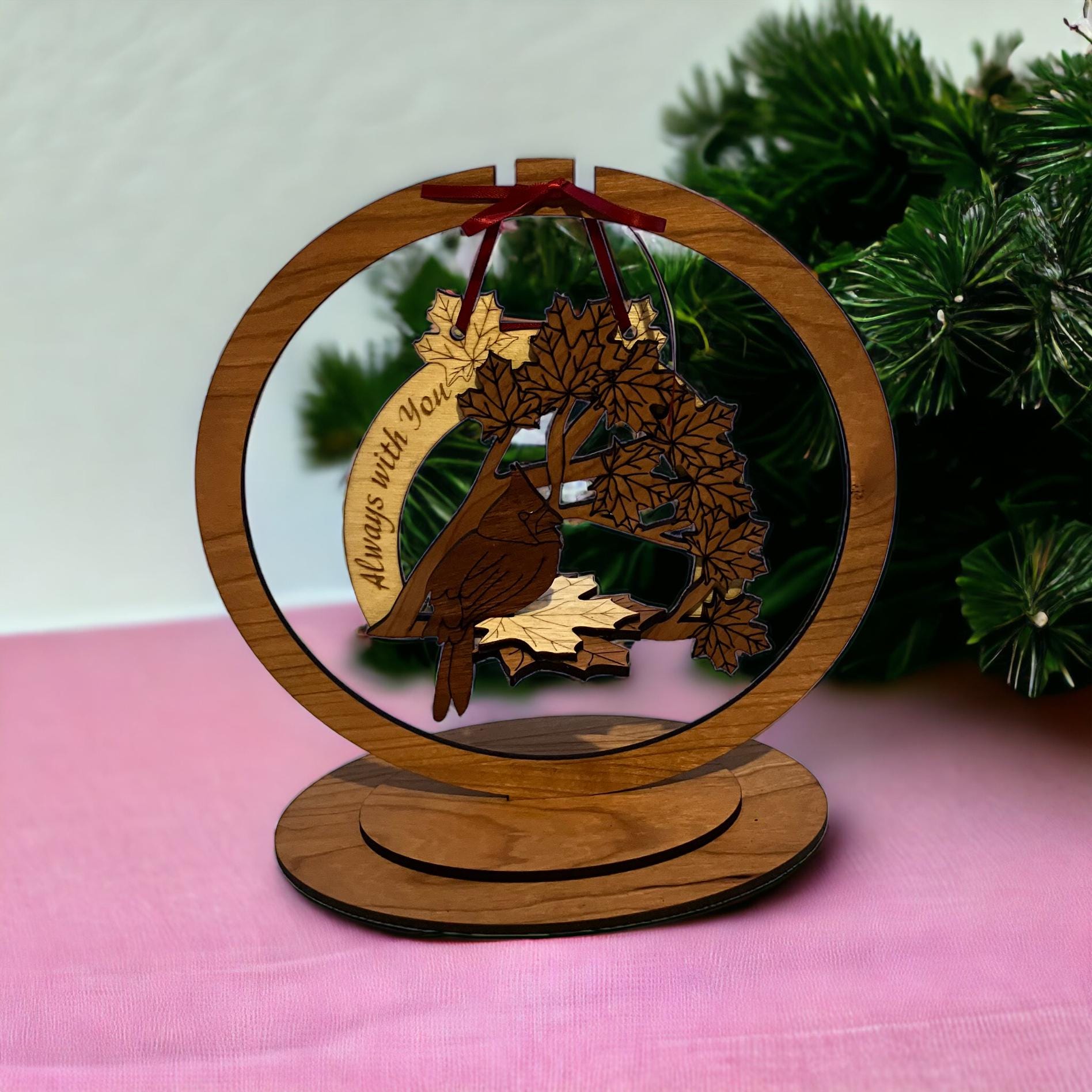 Wooden ornament with deer design and 'Always with you' text, placed on a pink surface with greenery in the background.