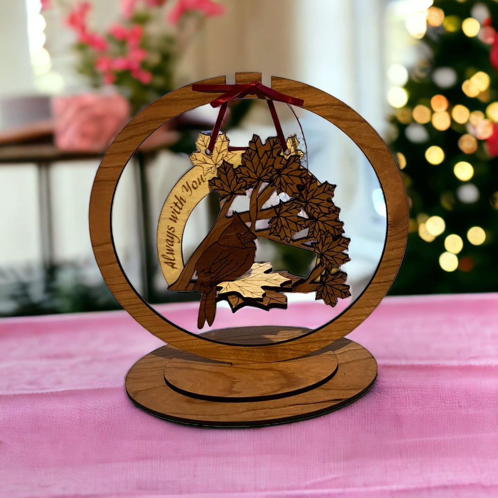 Wooden ornament with floral design and 'Always with you' text on a pink surface with blurred Christmas tree in the background.