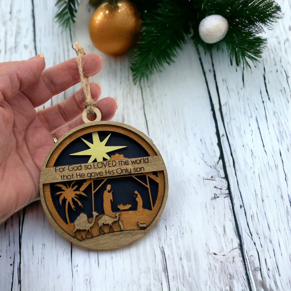 John 3:16 nativity ornament displayed on rustic wood background with greenery.